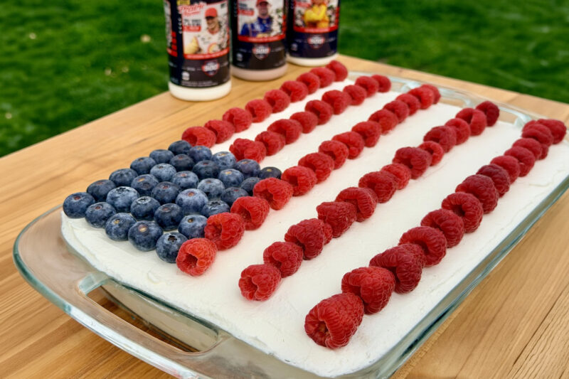 Rectangular ice cream cake topped with blueberries and raspberries arranged like an American flag, set in a glass dish with milk bottles in the background.