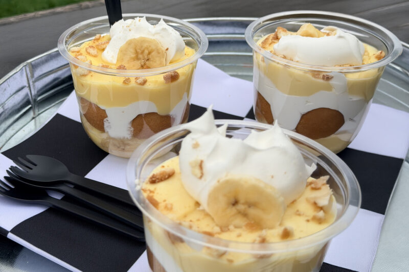 Three banana pudding cups layered with vanilla pudding, whipped cream, and cookies, topped with banana slices and cookie crumbles, served with plastic spoons on a tray.