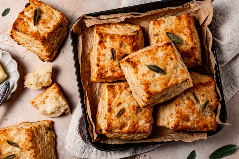 Golden, flaky buttermilk sage biscuits stacked in a parchment-lined pan with butter and sage leaves on the side.