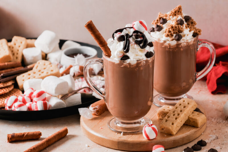 Two mugs of hot chocolate topped with whipped cream and assorted toppings, served beside a board of marshmallows, peppermint candies, cookies, and wafer sticks.