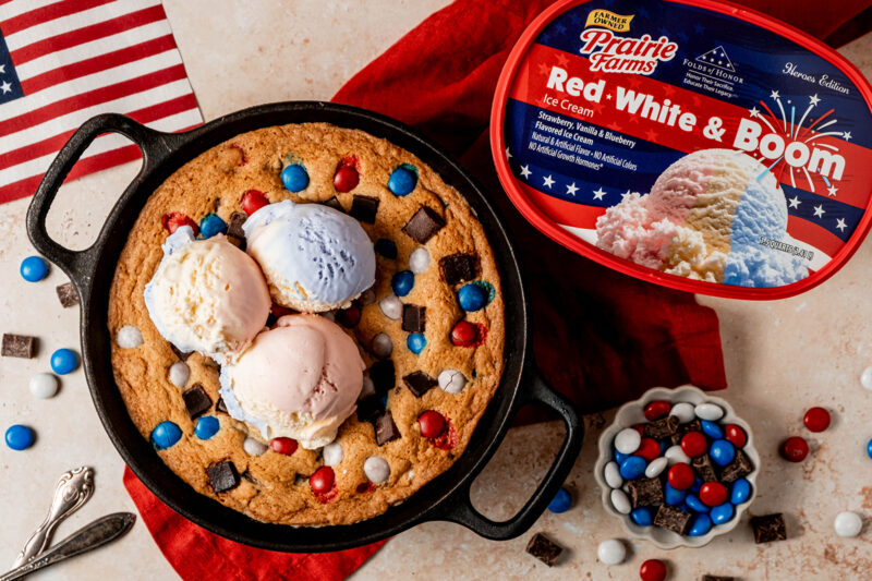 A patriotic skillet cookie topped with scoops of red, white, and blue ice cream, surrounded by colorful candy pieces and chocolate chunks, served in a cast iron skillet beside a container of Prairie Farms Red, White & Boom ice cream.