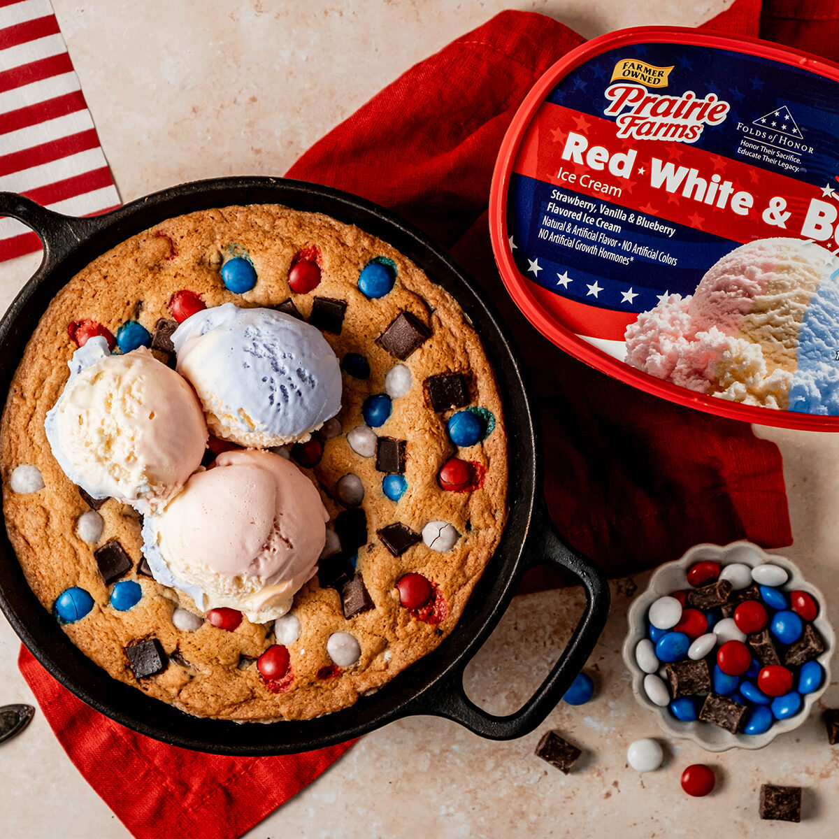 A patriotic skillet cookie topped with scoops of red, white, and blue ice cream, surrounded by colorful candy pieces and chocolate chunks, served in a cast iron skillet beside a container of Prairie Farms Red, White & Boom ice cream.