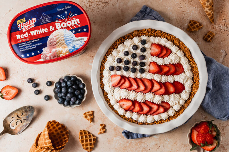 A patriotic ice cream pie decorated with whipped cream, strawberries, and blueberries in an American flag design, served in a waffle cone crust beside a container of Prairie Farms Red, White & Boom Ice Cream.