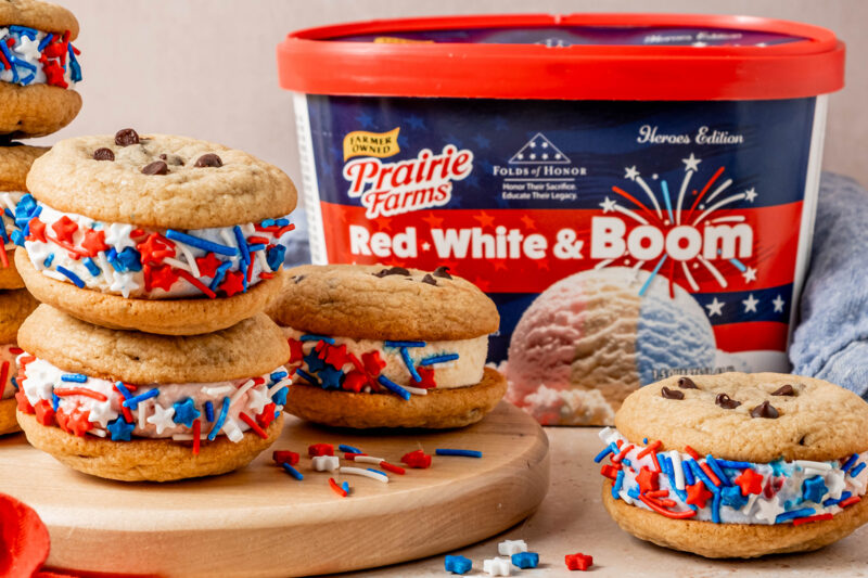 Chocolate chip cookie ice cream sandwiches filled with red, white, and blue ice cream and coated in patriotic sprinkles, arranged on a wooden board beside a container of Prairie Farms Red, White & Boom Ice Cream.