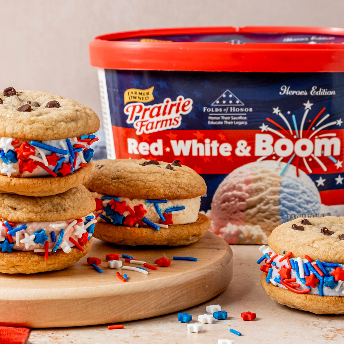 Chocolate chip cookie ice cream sandwiches filled with red, white, and blue ice cream and coated in patriotic sprinkles, arranged on a wooden board beside a container of Prairie Farms Red, White & Boom Ice Cream.
