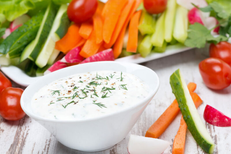 White bowl of creamy herb dip surrounded by sliced vegetables and cherry tomatoes